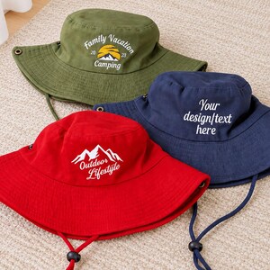 Puede incluir: Tres sombreros de pescador en verde oliva, azul marino y rojo. El sombrero oliva presenta un gráfico de sol y montaña amarillo con el texto Family Vacation 2023 Camping. El sombrero azul marino muestra el texto Your design/text here. El sombrero rojo tiene un gráfico de montaña blanco con el texto Outdoor Lifestyle.