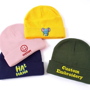 Peut inclure: Quatre bonnets en tricot de couleurs jaune, rose, bleu marine et vert olive. Le bonnet jaune présente un éléphant. Le bonnet rose a un visage souriant et le mot "CUSTOM". Le bonnet bleu marine a le texte "HA HA". Le bonnet vert olive dit "Custom Embroidery".