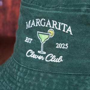 Pu&ograve; includere: Un cappello da pescatore verde scuro con testo ricamato bianco e un disegno di bicchiere da margarita. Il testo recita "Margarita Clever Club EST 2025". Il cappello &egrave; realizzato in un tessuto morbido e strutturato.
