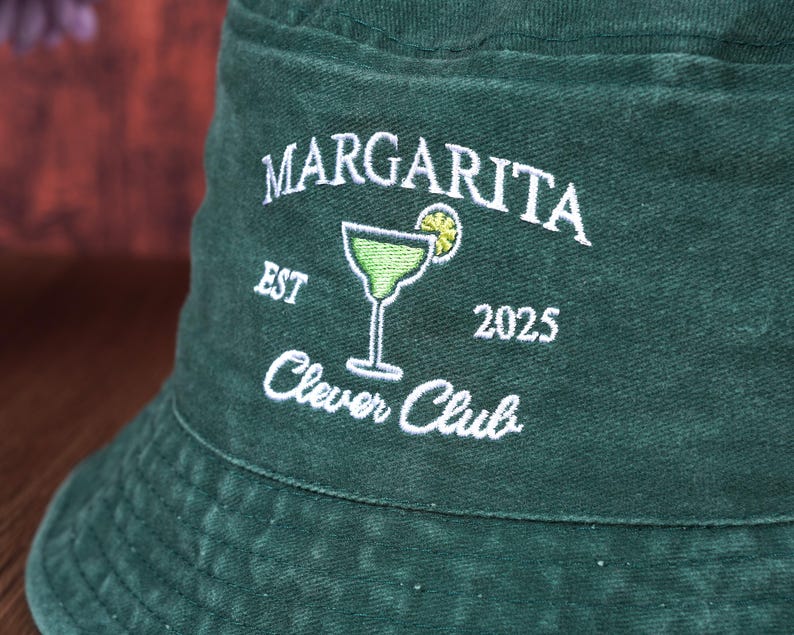 Pu&ograve; includere: Un cappello da pescatore verde scuro con testo ricamato bianco e un disegno di bicchiere da margarita. Il testo recita "Margarita Clever Club EST 2025". Il cappello &egrave; realizzato in un tessuto morbido e strutturato.