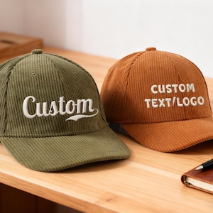 Puede incluir: Dos gorras de béisbol de pana. Una es verde oliva con "Custom" bordado en blanco, la otra es de color óxido con "CUSTOM TEXT/LOGO" bordado en blanco. Están sobre una superficie de madera.