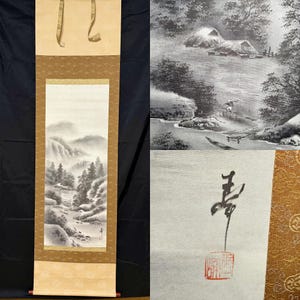 May include: A traditional Japanese scroll painting featuring a landscape scene with mountains, trees, and a river. The artwork is framed with a gold border and has a cream-colored top and bottom. The scroll is hanging against a black background.