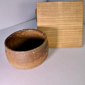 以下が含まれることがあります： 茶色とベージュの斑点模様がある、丸くて質感のある陶器のボウル。表面はざらざらとした土のような質感です。ボウルの横には、木目がはっきりと見える正方形で平らな薄茶色の木製の蓋が置かれています。