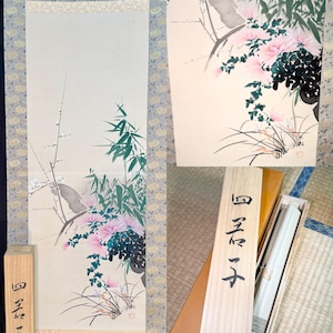 May include: A vertical hanging scroll featuring a painting of pink chrysanthemums, green bamboo, and plum blossoms on a light background. The scroll is framed by a patterned blue border and includes a wooden storage box with black Japanese calligraphy.