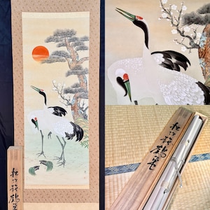 May include: A vertical hanging scroll painting featuring two white cranes with red crowns, a large orange sun, a pine tree, and white blossoms. The scroll is accompanied by a wooden storage box with black Japanese calligraphy characters on the lid.