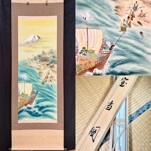 May include: A vertical hanging scroll featuring a detailed painting of a ship carrying figures towards a shoreline with Mount Fuji in the background. The scene includes a white crane, blue waves, and a wooden storage box with Japanese calligraphy.