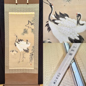 May include: A vertical hanging scroll featuring a painting of three white cranes with black wing tips and red crowns near a pine tree. The scroll has brown and patterned fabric borders. A wooden storage box with Japanese text is included.