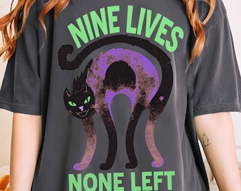May include: Dark gray t-shirt with the text "NINE LIVES NONE LEFT" in green and a black cat graphic with purple and green accents. The shirt is a comfortable, casual style.