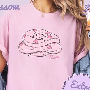 May include: A light pink t-shirt featuring a cartoon snake design with pink heart accents. The snake is coiled and has a smiling face. The word "Love" is printed below the snake. The shirt also has the word "Blossom" printed on the top left.