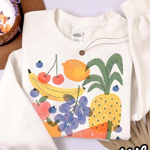 May include: White sweatshirt with a colorful fruit graphic including a banana, cherries, lemon, pineapple, grapes, and watermelon. The word "White" is written in a script font at the bottom right.