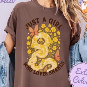 May include: Espresso-colored t-shirt with the text "JUST A GIRL WHO LOVES SNAKES." The graphic features a yellow snake surrounded by yellow flowers and butterflies. The shirt has a relaxed fit and short sleeves.