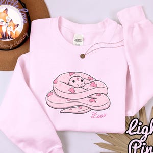 May include: Light pink sweatshirt featuring a cartoon snake design with pink heart accents. The word "Love" is printed below the snake. The phrase "light Pink" is written in a script font on the bottom right.