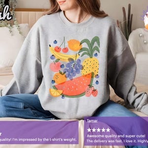 May include: A light gray sweatshirt featuring a colorful fruit design. The graphic includes a watermelon, pineapple, banana, grapes, oranges, strawberries, and cherries. The word "Ash" is in the top left corner.