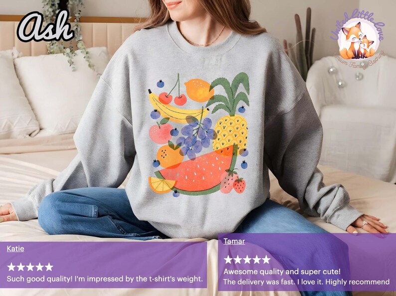 May include: A light gray sweatshirt featuring a colorful fruit design. The graphic includes a watermelon, pineapple, banana, grapes, oranges, strawberries, and cherries. The word "Ash" is in the top left corner.