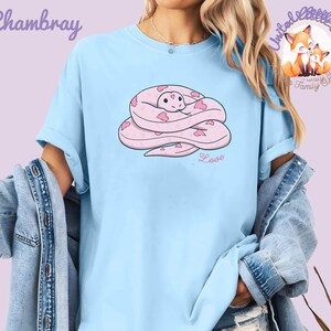 May include: Light blue t-shirt featuring a pink cartoon snake with heart-shaped markings. The word "Love" is written below the snake. The shirt also has the word "Chambray" and a logo with two foxes.
