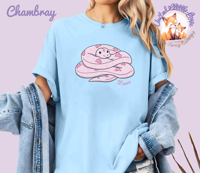 May include: Light blue t-shirt featuring a pink cartoon snake with heart-shaped markings. The word "Love" is written below the snake. The shirt also has the word "Chambray" and a logo with two foxes.