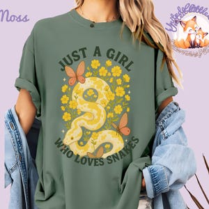 Pu&ograve; includere: T-shirt verde muschio con la scritta "JUST A GIRL WHO LOVES SNAKES". La grafica presenta un serpente giallo, fiori gialli e farfalle arancioni. La maglietta ha le maniche arrotolate.