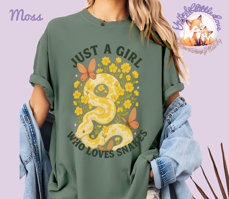 Pu&ograve; includere: T-shirt verde muschio con la scritta "JUST A GIRL WHO LOVES SNAKES". La grafica presenta un serpente giallo, fiori gialli e farfalle arancioni. La maglietta ha le maniche arrotolate.