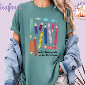 Peut inclure: T-shirt vert d'eau avec un motif de crayons color&eacute;s et le texte "color the world with kindness". Les crayons sont de diff&eacute;rentes couleurs, dont le bleu, le rose, le jaune et l'orange. Le t-shirt a des manches courtes et une coupe d&eacute;contract&eacute;e.