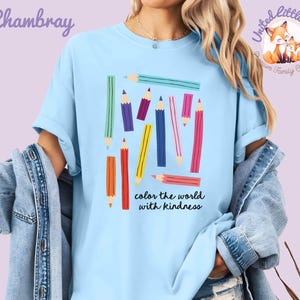 Peut inclure: T-shirt bleu clair avec un motif de crayons de couleur. Les crayons sont de diff&eacute;rentes couleurs, notamment rouge, jaune, bleu et violet. Le texte "color the world with kindness" est en dessous. Le t-shirt a des manches retrouss&eacute;es.