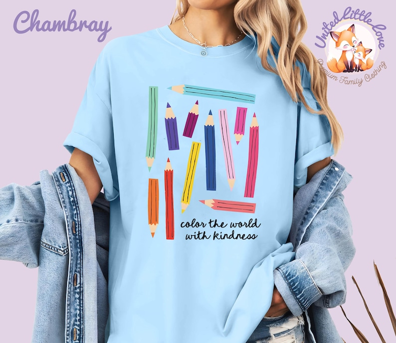 Peut inclure: T-shirt bleu clair avec un motif de crayons de couleur. Les crayons sont de diff&eacute;rentes couleurs, notamment rouge, jaune, bleu et violet. Le texte "color the world with kindness" est en dessous. Le t-shirt a des manches retrouss&eacute;es.