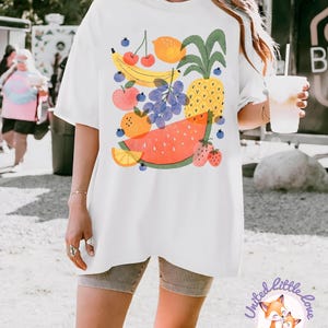 May include: White t-shirt featuring a colorful fruit illustration. The design includes a banana, cherries, lemon, pineapple, grapes, apple, orange, watermelon, and strawberries. The shirt has a relaxed fit and short sleeves. The logo "United Little Love" is at the bottom.