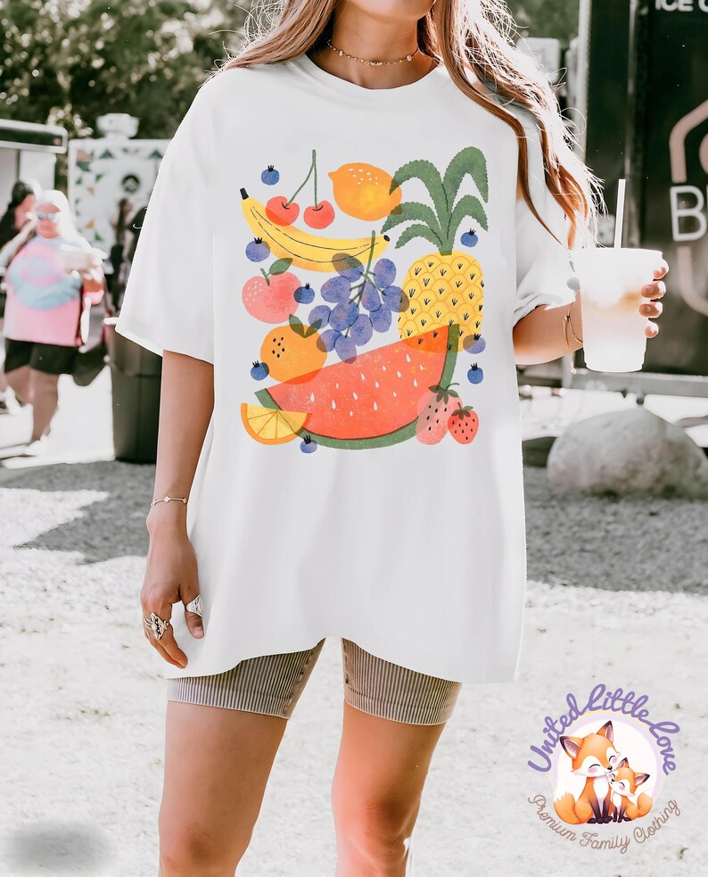 May include: White t-shirt featuring a colorful fruit illustration. The design includes a banana, cherries, lemon, pineapple, grapes, apple, orange, watermelon, and strawberries. The shirt has a relaxed fit and short sleeves. The logo "United Little Love" is at the bottom.