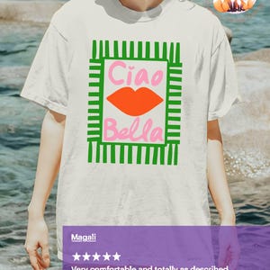 K&ouml;nnte beinhalten: Wei&szlig;es T-Shirt mit einem gr&uuml;n-rosa Grafikdesign. Das Design zeigt die Worte "Ciao Bella" in Pink, mit einer orangefarbenen Lippen-Grafik. Das T-Shirt ist ein l&auml;ssiger Stil.