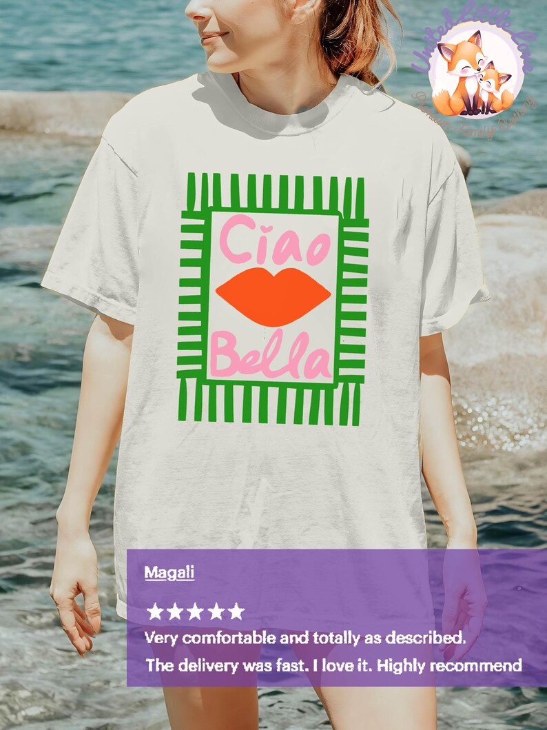 K&ouml;nnte beinhalten: Wei&szlig;es T-Shirt mit einem gr&uuml;n-rosa Grafikdesign. Das Design zeigt die Worte "Ciao Bella" in Pink, mit einer orangefarbenen Lippen-Grafik. Das T-Shirt ist ein l&auml;ssiger Stil.