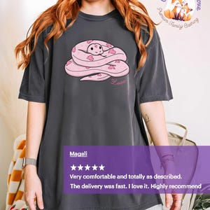 May include: Dark gray t-shirt featuring a pink snake design with heart accents and the word "Love". The shirt has short sleeves and a relaxed fit. The brand name "Pepper" is in the upper left corner.