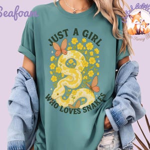 Pu&ograve; includere: T-shirt verde acqua con la scritta "JUST A GIRL WHO LOVES SNAKES". La grafica presenta un serpente giallo con accenti floreali e farfalle. La maglietta &egrave; uno stile comodo e casual.