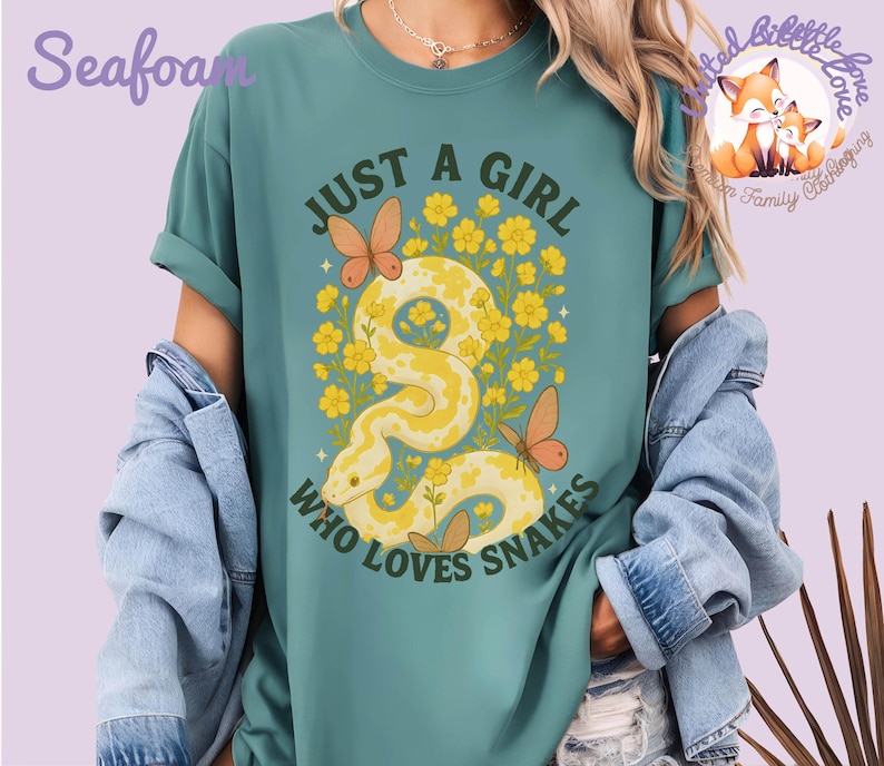 Pu&ograve; includere: T-shirt verde acqua con la scritta "JUST A GIRL WHO LOVES SNAKES". La grafica presenta un serpente giallo con accenti floreali e farfalle. La maglietta &egrave; uno stile comodo e casual.