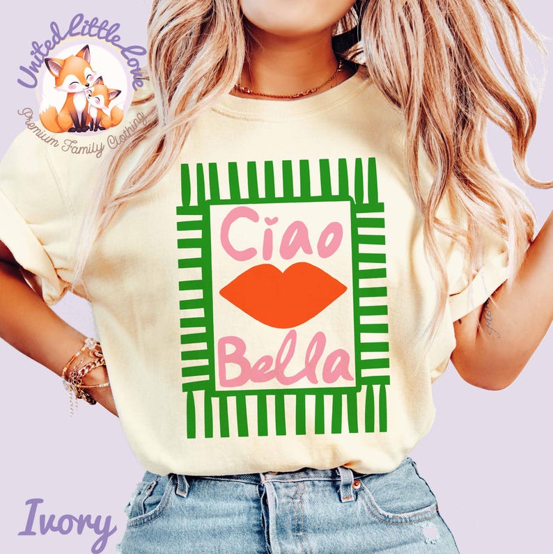 K&ouml;nnte beinhalten: Elfenbeinfarbenes T-Shirt mit einem gr&uuml;n-wei&szlig;en rechteckigen Design. Das Design enth&auml;lt die Worte "Ciao Bella" in Rosa und eine orangefarbene Lippen-Grafik. Das T-Shirt hat kurze &Auml;rmel und ist aus weichem Material. Teil der "Premium Family Clothing"-Linie.