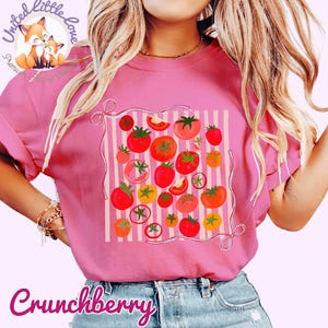 Peut inclure: T-shirt rose avec un motif de tomates color&eacute;es. Le design pr&eacute;sente diverses tomates rouges, oranges et jaunes, dispos&eacute;es sur un fond ray&eacute;. Le mot "Crunchberry" est &eacute;crit en cursive en bas.