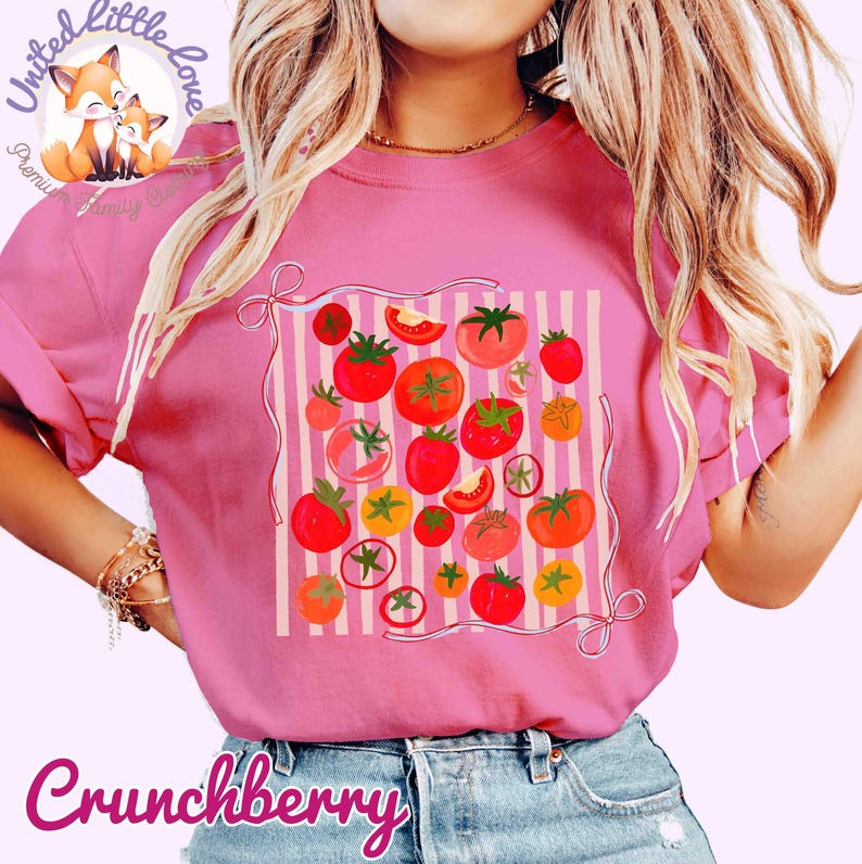 Peut inclure: T-shirt rose avec un motif de tomates color&eacute;es. Le design pr&eacute;sente diverses tomates rouges, oranges et jaunes, dispos&eacute;es sur un fond ray&eacute;. Le mot "Crunchberry" est &eacute;crit en cursive en bas.