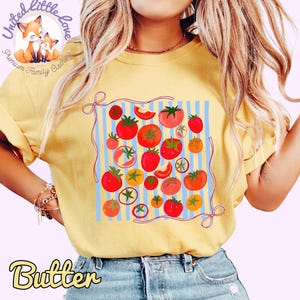 Peut inclure: T-shirt jaune avec un motif de tomates. Le design pr&eacute;sente diverses tomates rouges et oranges avec des tiges vertes, sur un fond &agrave; rayures bleues. Le mot "Butter" est imprim&eacute; en bas.