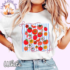 Peut inclure: T-shirt blanc avec un motif de tomates sur un fond &agrave; rayures bleues et blanches. Le motif comprend des tomates rouges et oranges de diff&eacute;rentes tailles, avec une bordure en ruban rouge. Le mot "White" est en bas.