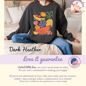 May include: Dark heather sweatshirt featuring a colorful fruit illustration. The design includes a pineapple, watermelon, grapes, strawberries, and other fruits. The text "love it guarantee" is below the sweatshirt.