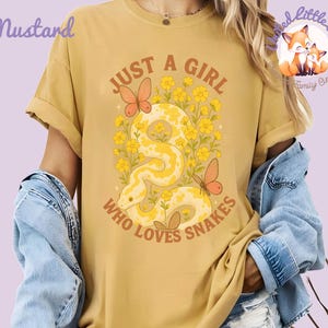 Pu&ograve; includere: T-shirt color senape con grafica di un serpente giallo, farfalle e fiori. Il testo recita "JUST A GIRL WHO LOVES SNAKES". La maglietta ha le maniche arrotolate e fa parte della collezione "Little Love Family Clothing".