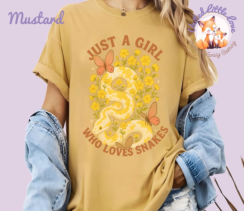 Pu&ograve; includere: T-shirt color senape con grafica di un serpente giallo, farfalle e fiori. Il testo recita "JUST A GIRL WHO LOVES SNAKES". La maglietta ha le maniche arrotolate e fa parte della collezione "Little Love Family Clothing".