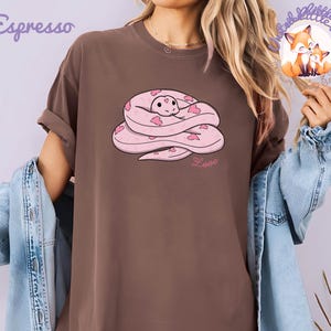 May include: Espresso-colored t-shirt featuring a cartoon snake design. The snake is pink with heart-shaped markings and a smiling face. The word "Love" is written below the snake in pink script. The shirt has a relaxed fit.
