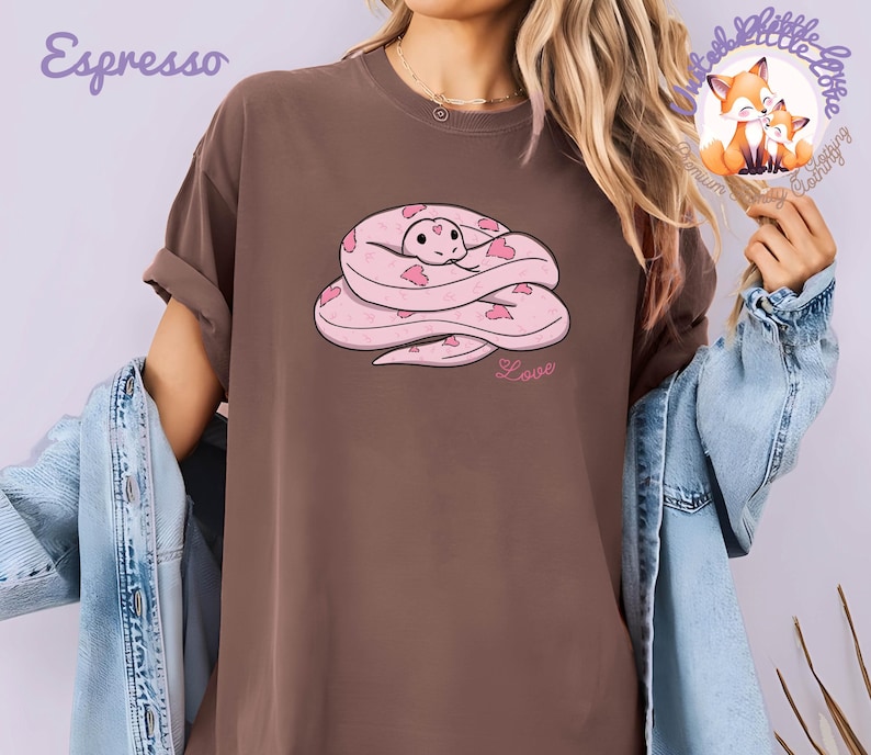 May include: Espresso-colored t-shirt featuring a cartoon snake design. The snake is pink with heart-shaped markings and a smiling face. The word "Love" is written below the snake in pink script. The shirt has a relaxed fit.