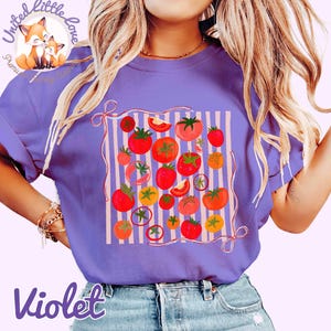 Peut inclure: T-shirt violet avec un motif de tomates. Le motif pr&eacute;sente des tomates rouges, oranges et jaunes sur un fond ray&eacute;. Le t-shirt a des manches courtes et un col rond. Le texte "Violet" est en bas du t-shirt.