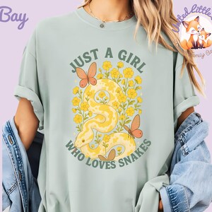 Pu&ograve; includere: T-shirt verde chiaro con la scritta "JUST A GIRL WHO LOVES SNAKES" in verde. La grafica presenta un serpente giallo, fiori gialli e farfalle arancioni. La maglietta &egrave; in stile casual.