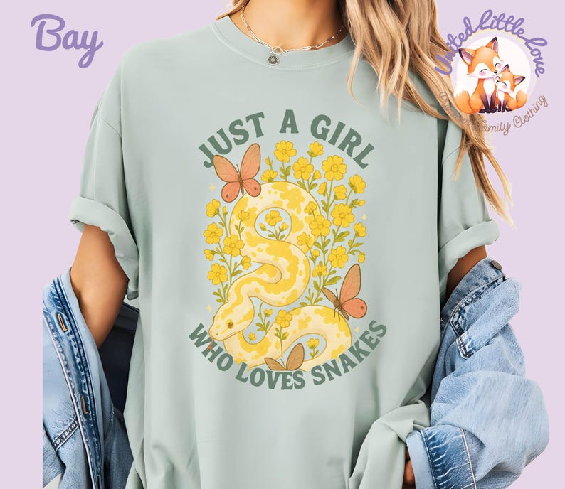 Pu&ograve; includere: T-shirt verde chiaro con la scritta "JUST A GIRL WHO LOVES SNAKES" in verde. La grafica presenta un serpente giallo, fiori gialli e farfalle arancioni. La maglietta &egrave; in stile casual.