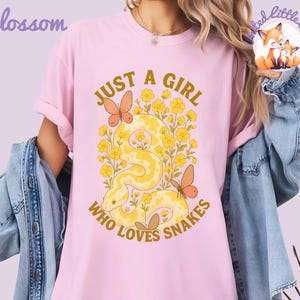 Pu&ograve; includere: T-shirt rosa chiaro con la scritta dorata "JUST A GIRL WHO LOVES SNAKES". Il design presenta un serpente giallo, fiori gialli e farfalle arancioni. Le maniche sono arrotolate.