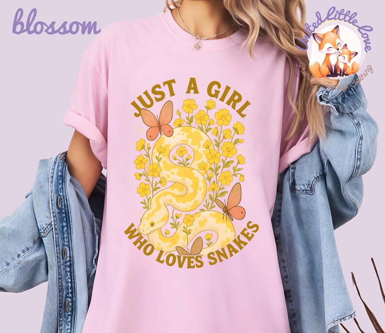 Pu&ograve; includere: T-shirt rosa chiaro con la scritta dorata "JUST A GIRL WHO LOVES SNAKES". Il design presenta un serpente giallo, fiori gialli e farfalle arancioni. Le maniche sono arrotolate.