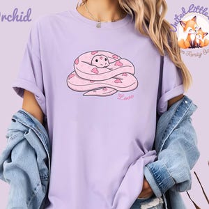 May include: A light purple t-shirt with a cartoon snake design. The snake is pink with heart-shaped markings and is coiled up. The word "Love" is written in pink script below the snake. The word "Orchid" is printed in purple at the top left.