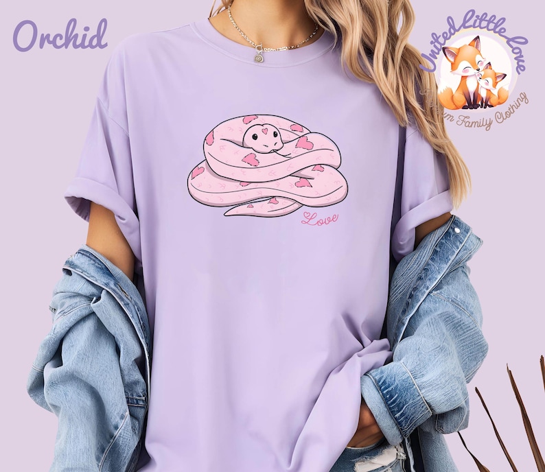 May include: A light purple t-shirt with a cartoon snake design. The snake is pink with heart-shaped markings and is coiled up. The word "Love" is written in pink script below the snake. The word "Orchid" is printed in purple at the top left.