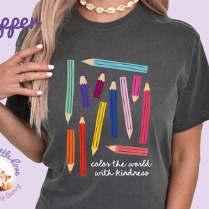 Peut inclure: T-shirt gris fonc&eacute; avec un motif de crayons color&eacute;s. Les crayons sont de diff&eacute;rentes couleurs, notamment bleu, vert, violet, jaune et rose. Le texte "color the world with kindness" est imprim&eacute; en dessous.